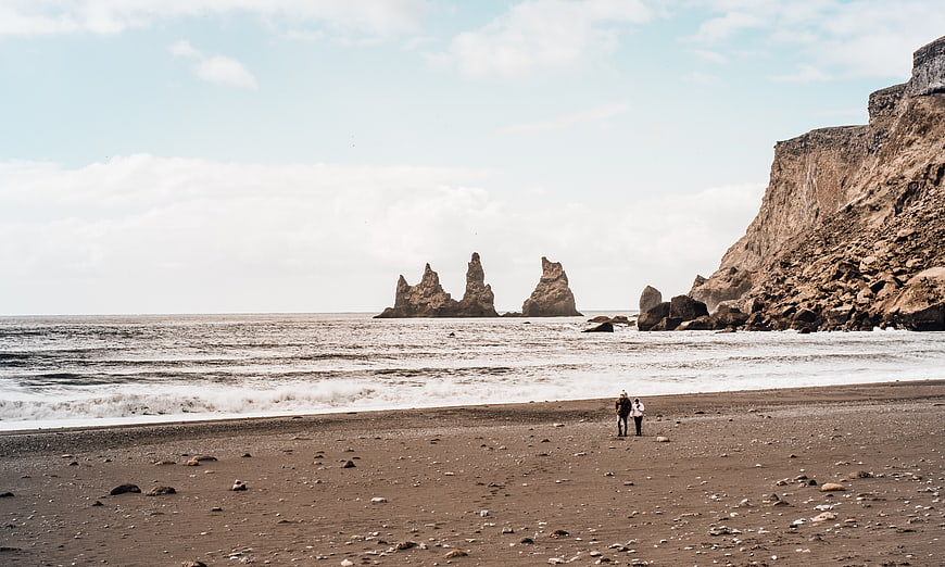 Couple walking on black sand beach with sea stacks