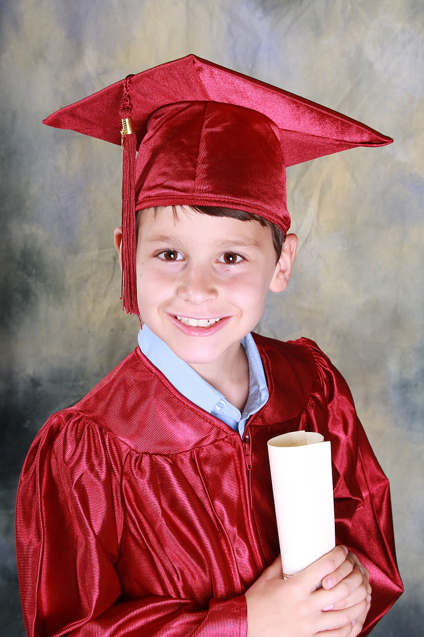 Happy young boy in red graduation cap and gown holding diploma