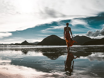 Woman in Orange Dress Walking on Wet Beach with Mountain Reflection