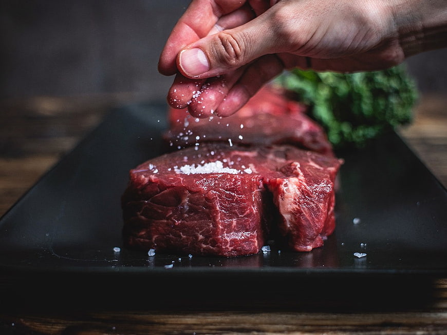 Hand Seasoning Raw Steak with Salt on Black Plate