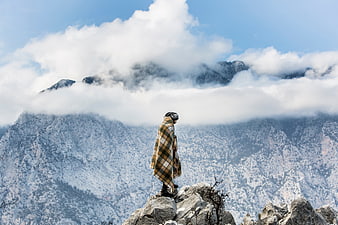 Man in plaid blanket standing on rocky mountain peak with clouds