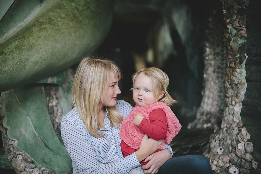 Mother holding toddler daughter in front of mossy stone statue