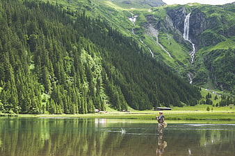 Man fly fishing in alpine lake with waterfall and forest