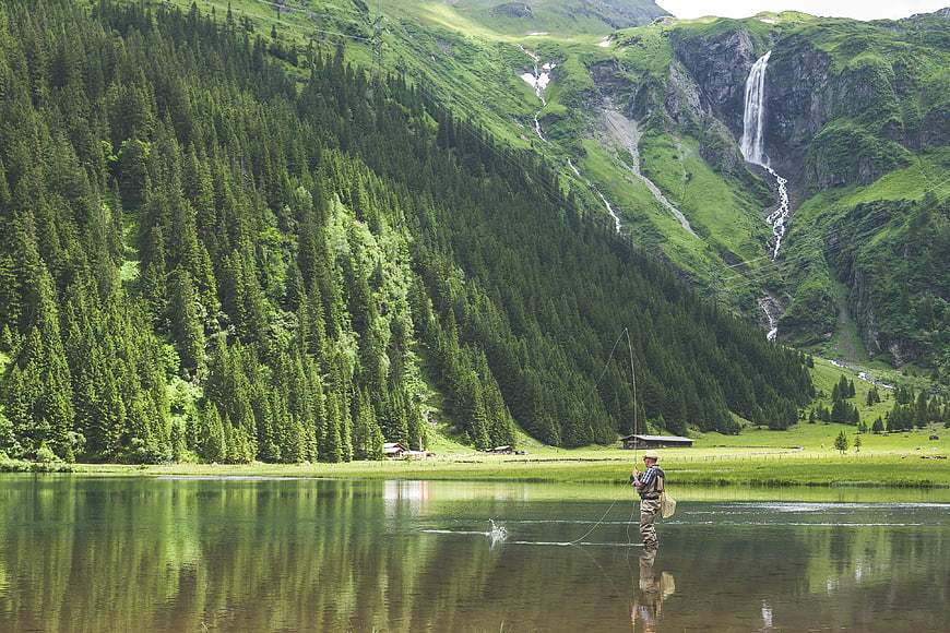 Man fly fishing in alpine lake with waterfall and forest