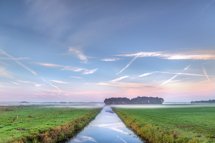 Misty Canal Through Green Meadow at Dawn with Contrails