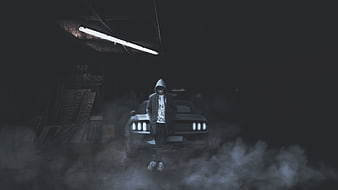 Man in Hoodie Standing in Front of Car in Smoky Garage