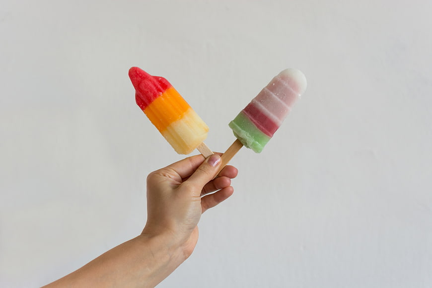 Hand holding two colorful striped popsicles on white background