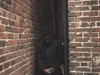 African American man in black suit checking watch against brick wall