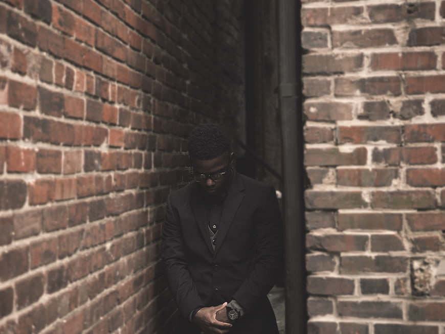 African American man in black suit checking watch against brick wall