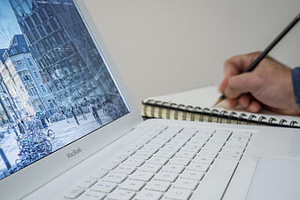 Hand writing in notebook next to white MacBook laptop