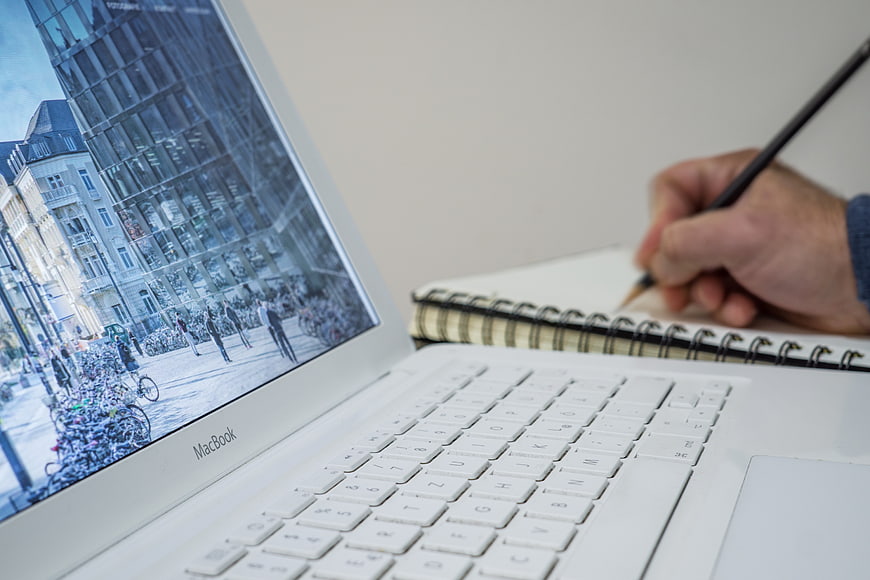 Hand writing in notebook next to white MacBook laptop