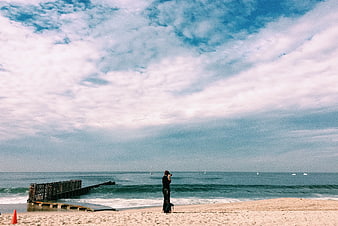 Person standing on sandy beach looking at ocean with pier