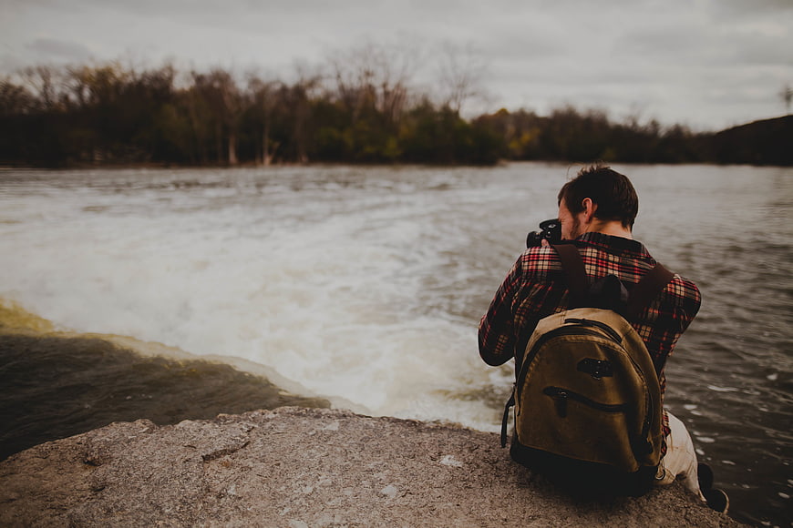 Photographer with Camera Sitting by Riverbank