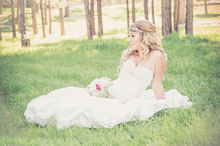 Bride in White Dress Sitting in Grassy Forest with Bouquet