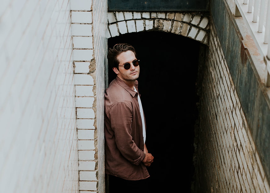 Stylish man in sunglasses leaning against white brick wall