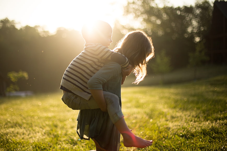 Siblings Piggyback Ride In Golden Hour Grass Field