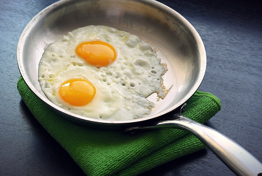Two sunny-side-up eggs in a stainless steel skillet on a green towel