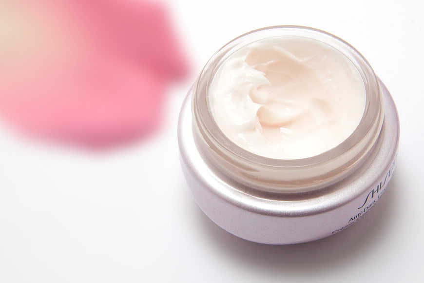 Open jar of white eye cream with blurred pink rose petal
