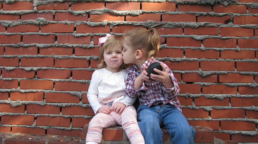 Two young girls sitting on a brick wall, one kissing the other on the cheek