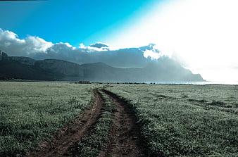 Dirt path leading through green field to mountains and sea