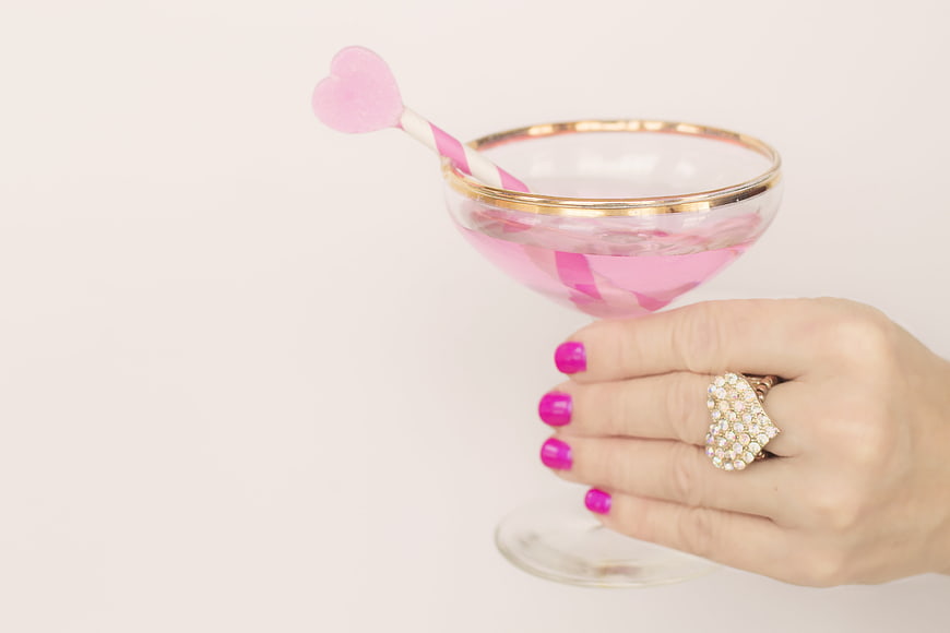 Hand holding gold-rimmed champagne glass with pink drink and heart stirrer