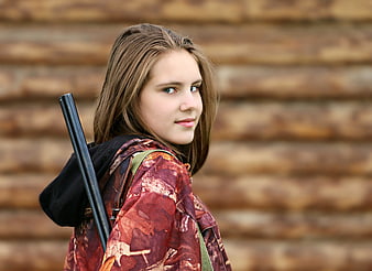 Young woman with rifle standing in front of rustic wooden log wall