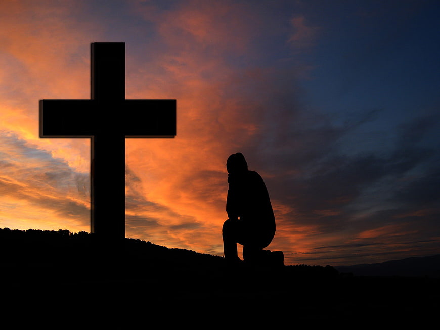 Silhouette of person kneeling in prayer by cross at sunset
