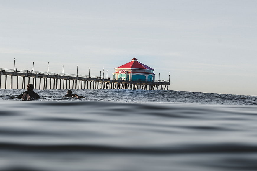 Surfers paddling in ocean with pier and red-roofed building in background