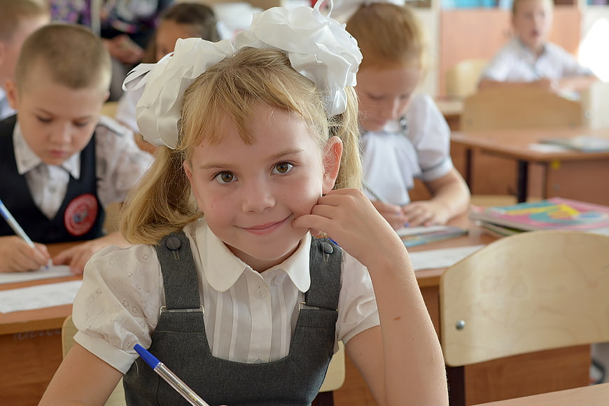 Smiling young schoolgirl with white hair bow sitting at a classroom desk