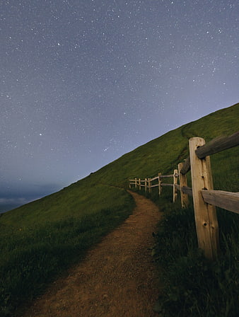 Dirt path along grassy hill with wooden fence under starry night sky