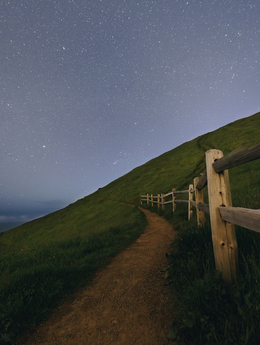 Dirt path along grassy hill with wooden fence under starry night sky
