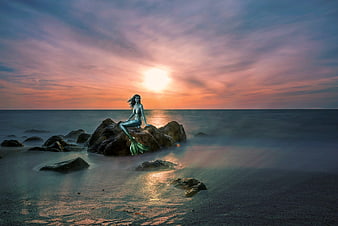 Fantasy mermaid sitting on rocks at sunset with long exposure ocean
