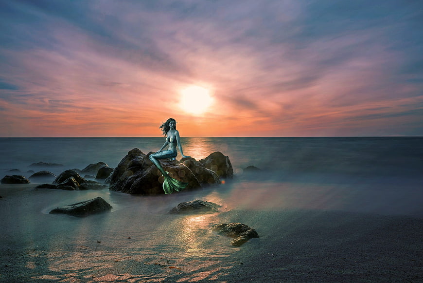 Fantasy mermaid sitting on rocks at sunset with long exposure ocean