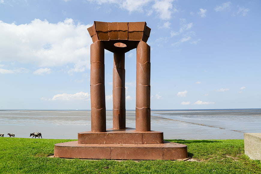 Rust-colored abstract sculpture on grassy coast with sea and cows