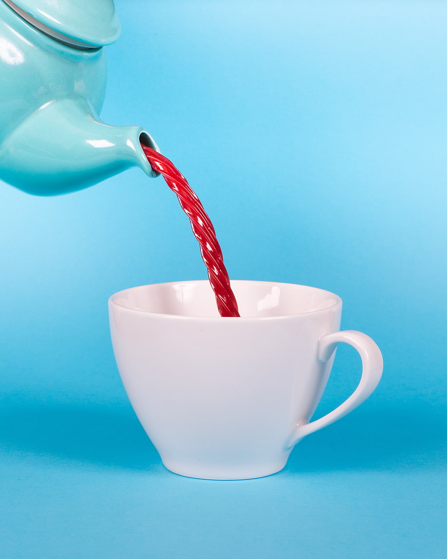 Red liquid pouring from turquoise teapot into white cup on blue background