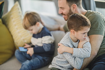 Sad boy resting on father's lap while brother plays with phone
