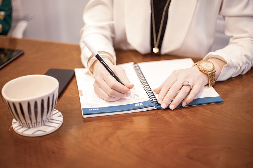 Woman writing in notebook at wooden desk with coffee cup