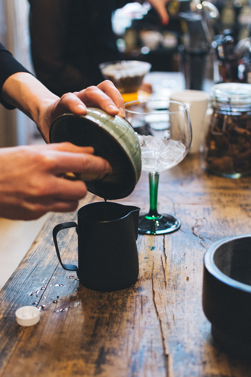 Hands pouring matcha tea into a black pitcher on a wooden table