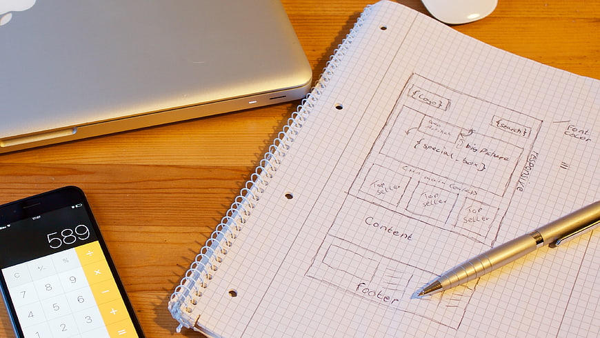Hand-drawn website wireframe sketch on grid notebook with laptop and calculator