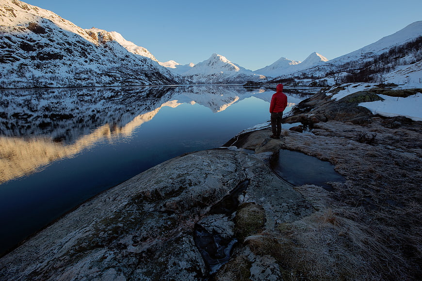 Man in Red Jacket Standing by Snowy Mountain Lake
