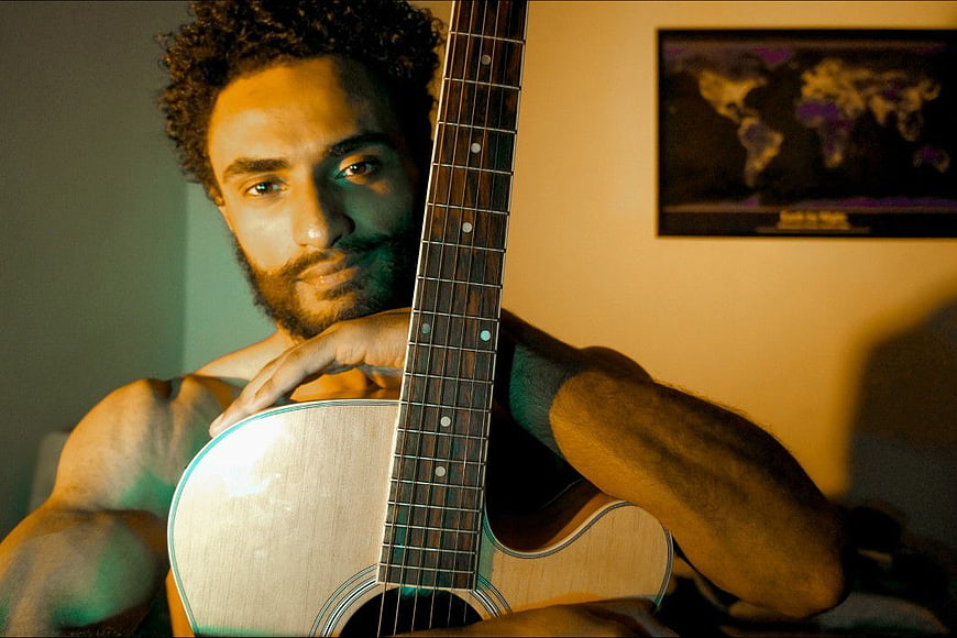 Shirtless male musician holding acoustic guitar with teal and orange lighting