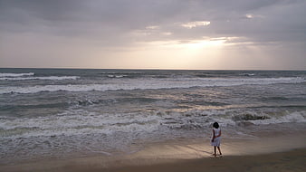 Woman standing on beach shore facing ocean waves at sunset