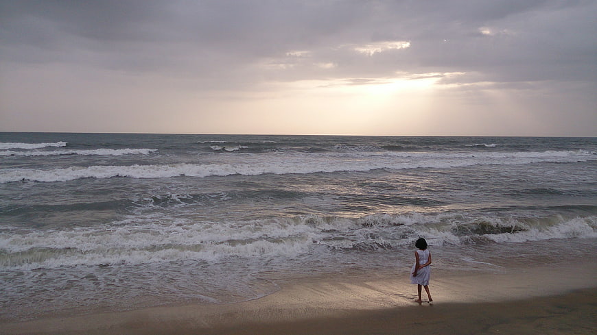 Woman standing on beach shore facing ocean waves at sunset