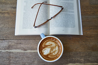 Flat lay of latte art coffee, open book, and glasses on wood