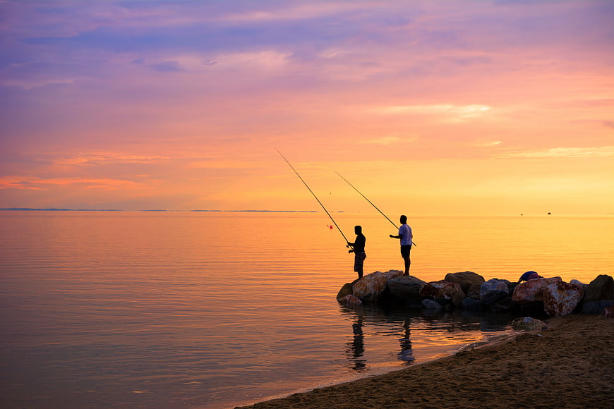 Two Silhouetted Fishermen on Rocks at Sunset Over Calm Sea