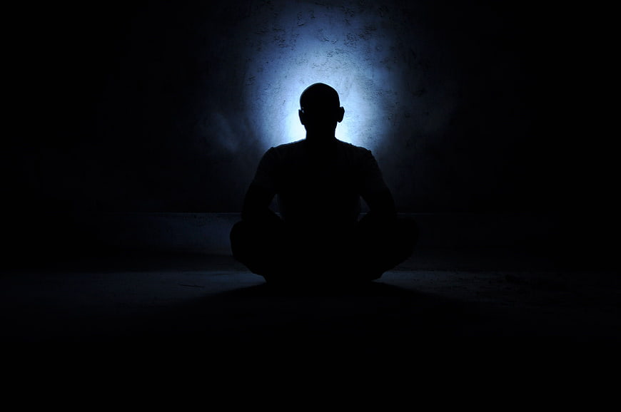 Silhouette of a person meditating in a dark room with a glowing blue aura