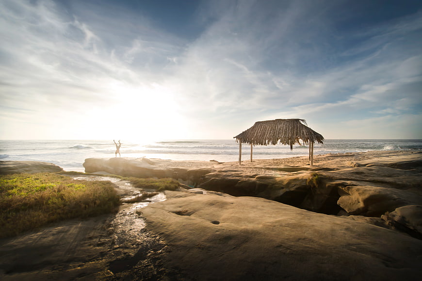 Man doing handstand on rocky beach with thatched hut at sunrise
