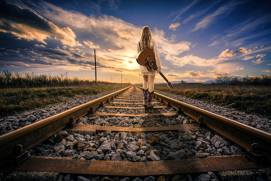 Woman with Guitar Walking on Railroad Tracks at Sunset