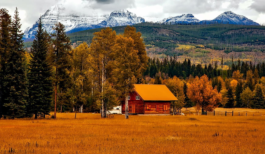 Red Log Cabin In Autumn Meadow With Snowy Mountains