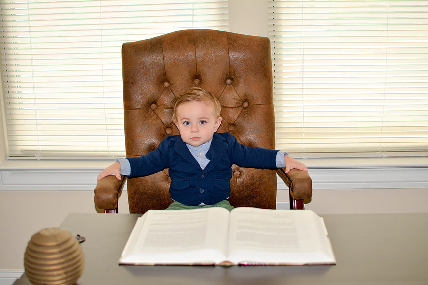 Toddler boy in navy blazer sitting in large leather office chair